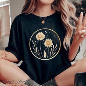 May include: Black heather t-shirt featuring a gold-framed floral design with two yellow sunflowers, leaves, and stars. The text "Black Heather" is visible at the bottom of the image. The shirt is a casual, comfortable style.