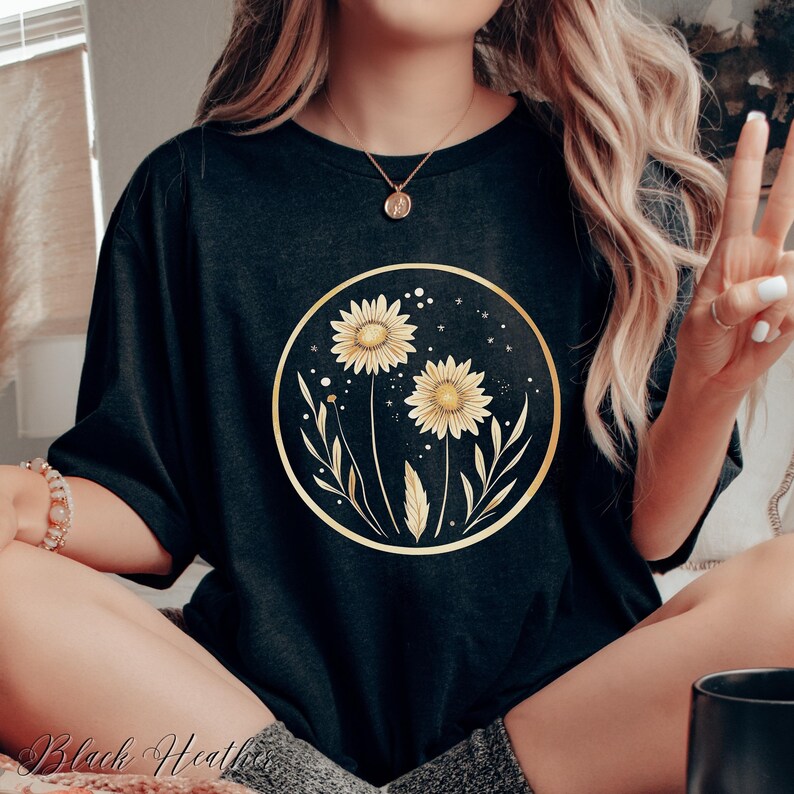 May include: Black heather t-shirt featuring a gold-framed floral design with two yellow sunflowers, leaves, and stars. The text "Black Heather" is visible at the bottom of the image. The shirt is a casual, comfortable style.
