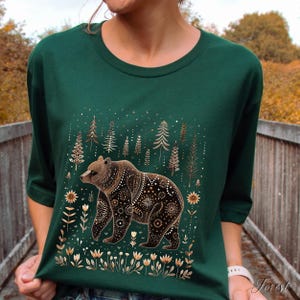 May include: A dark green t-shirt featuring a detailed illustration of a brown bear with decorative patterns. The bear is surrounded by stylized trees, flowers, and stars in shades of gold and white. The word "Forest" is printed on the bottom right.