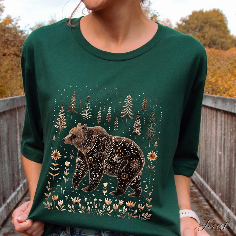 May include: A dark green t-shirt featuring a detailed illustration of a brown bear with decorative patterns. The bear is surrounded by stylized trees, flowers, and stars in shades of gold and white. The word "Forest" is printed on the bottom right.