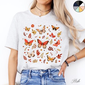 May include: A white t-shirt with a colorful butterfly and floral print. The butterflies are orange, red, and yellow with black outlines. The flowers are yellow, red, and pink. The shirt is a light grey color.