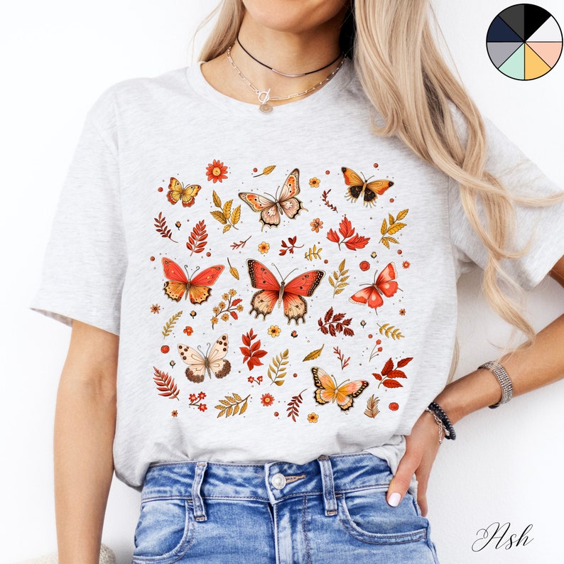May include: A white t-shirt with a colorful butterfly and floral print. The butterflies are orange, red, and yellow with black outlines. The flowers are yellow, red, and pink. The shirt is a light grey color.