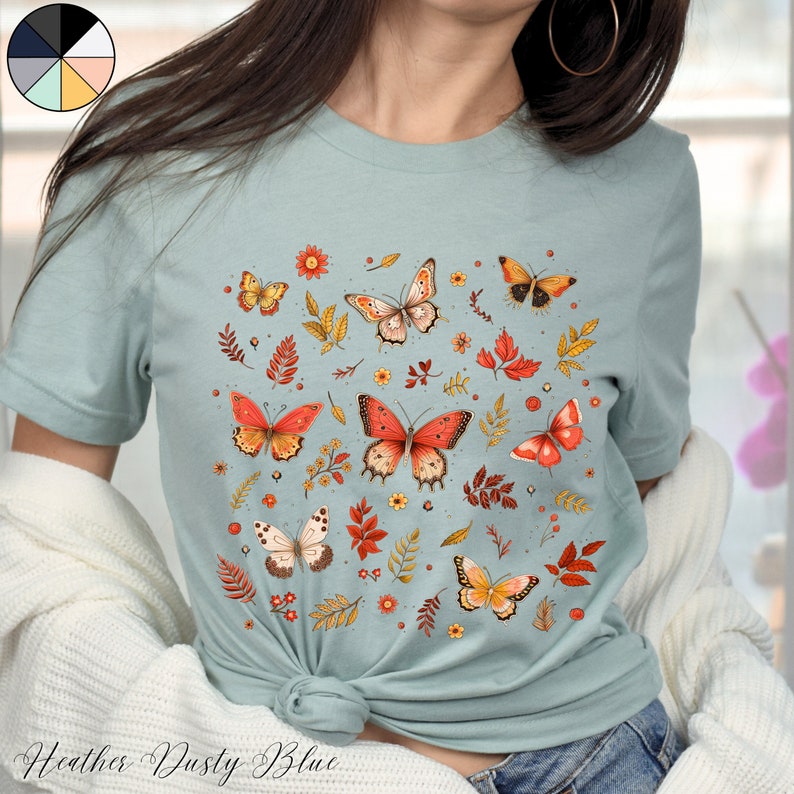 May include: A light blue t-shirt with a pattern of orange and yellow butterflies, leaves, and flowers. The text "Heather Dusty Blue" is printed on the bottom of the shirt.