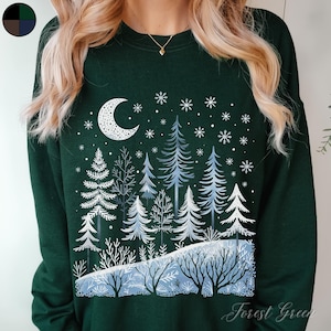 May include: A forest green sweatshirt with a white design of a crescent moon and a snowy forest scene. The text "Forest Green" is printed on the bottom right corner of the sweatshirt.