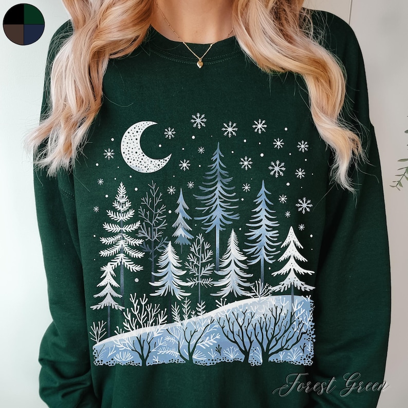 May include: A forest green sweatshirt with a white design of a crescent moon and a snowy forest scene. The text "Forest Green" is printed on the bottom right corner of the sweatshirt.