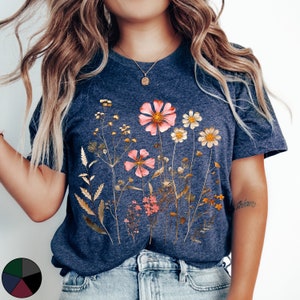 May include: A navy blue t-shirt with a floral print featuring pink, white, and yellow flowers. The flowers are arranged in a cluster and are surrounded by green leaves. The shirt has a crew neck and short sleeves.