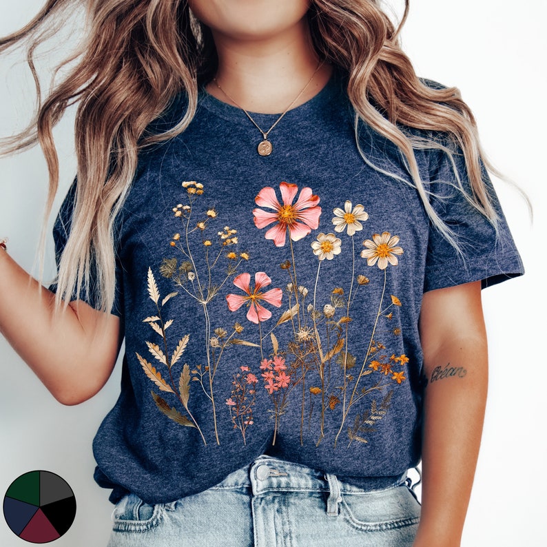 May include: A navy blue t-shirt with a floral print featuring pink, white, and yellow flowers. The flowers are arranged in a cluster and are surrounded by green leaves. The shirt has a crew neck and short sleeves.
