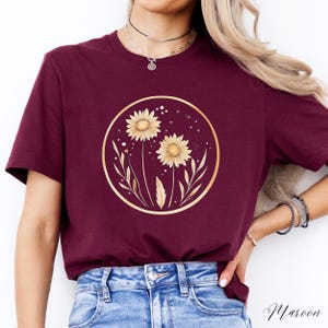 May include: Maroon-colored t-shirt featuring a gold-colored circular design with two sunflowers and botanical elements. The design is centered on the shirt. The word "Maroon" is written in script at the bottom right corner.