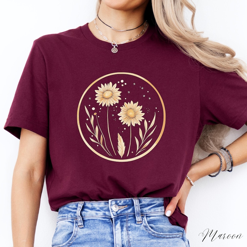 May include: Maroon-colored t-shirt featuring a gold-colored circular design with two sunflowers and botanical elements. The design is centered on the shirt. The word "Maroon" is written in script at the bottom right corner.
