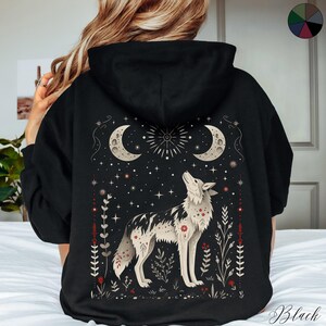 May include: Black hooded sweatshirt with a graphic print of a wolf howling at the moon. The design features a starry night sky with crescent moons and floral accents.