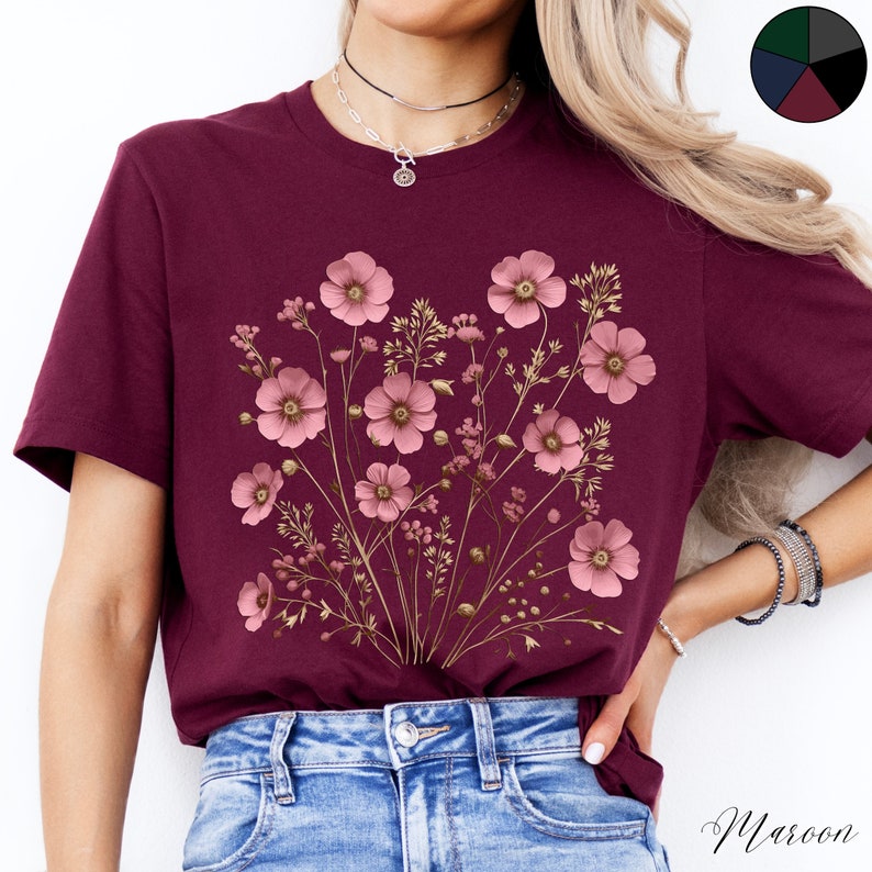 Pu&ograve; includere: Una t-shirt color granata con un design floreale che presenta fiori rosa e steli dorati. I fiori sono disposti a forma di bouquet e sono centrati sulla t-shirt.