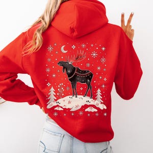 May include: A red hooded sweatshirt featuring a black moose design with antlers, standing on a snowy hill. The design includes snowflakes, a crescent moon, and decorative elements. The sweatshirt is a solid red color.