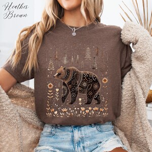 May include: Heather Brown t-shirt featuring a detailed bear illustration with floral and geometric patterns. The bear is surrounded by stylized trees and flowers in shades of brown and gold. The shirt is a neutral brown color.