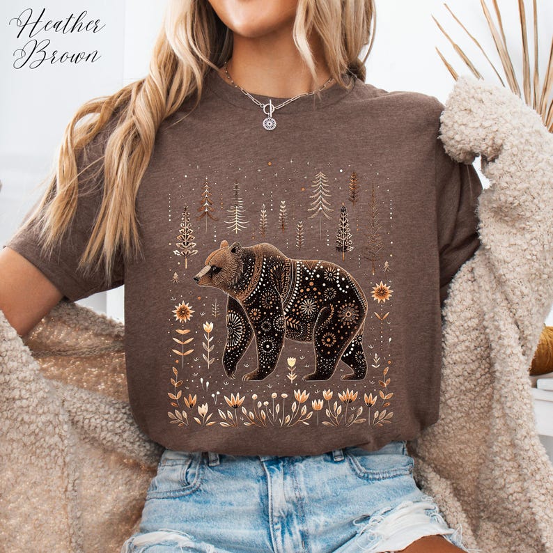 May include: Heather Brown t-shirt featuring a detailed bear illustration with floral and geometric patterns. The bear is surrounded by stylized trees and flowers in shades of brown and gold. The shirt is a neutral brown color.