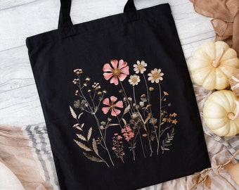 May include: Black tote bag with a floral design featuring pressed pink and white flowers on a black background. The flowers are arranged in a cluster and are surrounded by dried leaves and stems.