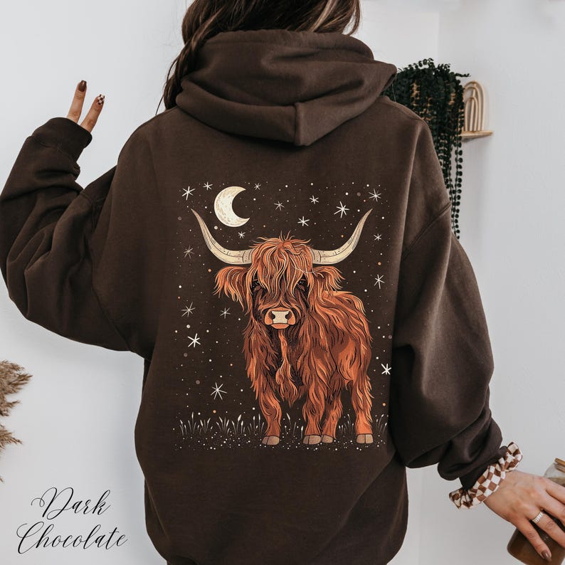 Op de afbeelding: Donker chocoladebruine hoodie met een Highland-koeienafbeelding op de rug. Het ontwerp bevat een halve maan, sterren en de tekst "Dark Chocolate". De koe is afgebeeld in bruin- en oranjetinten.