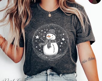 May include: A dark grey heather t-shirt with a white snowman graphic. The snowman is wearing a black hat and a scarf. The graphic is surrounded by white stars.
