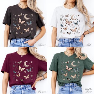 May include: Four different color t-shirts featuring a graphic print of butterflies, a crescent moon, and floral elements. The colors are brown, ash, maroon, and heather forest.