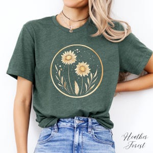 May include: A forest green t-shirt featuring a gold circular design with two yellow sunflowers and delicate leaves. The text "Heather Forest" is written in cursive on the bottom right.