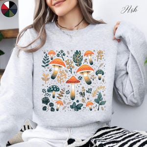 May include: A gray sweatshirt with a colorful mushroom and floral print. The print features orange, yellow, green, and white mushrooms with green leaves and flowers.