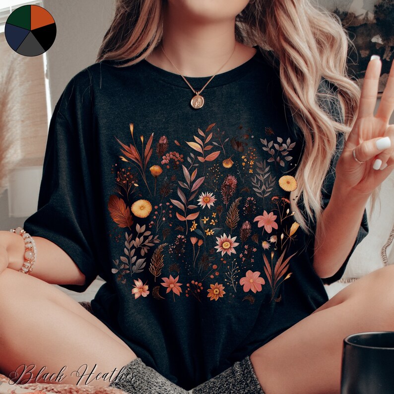 K&ouml;nnte beinhalten: Ein schwarzes T-Shirt mit einem Blumenmuster, das verschiedene Blumen und Bl&auml;tter in Orange-, Gelb- und Rosat&ouml;nen zeigt. Das Shirt hat eine lockere Passform und einen Rundhalsausschnitt.