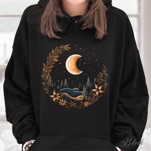 May include: Black hoodie with a gold and white crescent moon graphic. The moon is surrounded by a wreath of gold leaves and flowers. The graphic is set against a dark blue background with white stars.