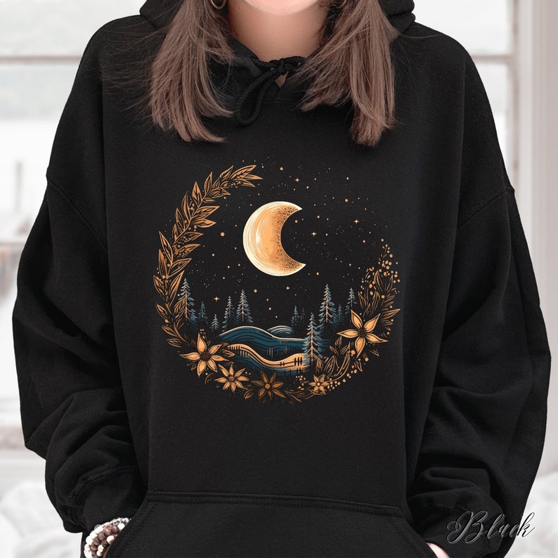 May include: Black hoodie with a gold and white crescent moon graphic. The moon is surrounded by a wreath of gold leaves and flowers. The graphic is set against a dark blue background with white stars.