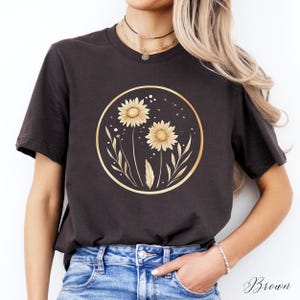 May include: Brown t-shirt featuring a gold circular design with two yellow sunflowers and leaves. The design is centered on the shirt. The word "Brown" is written in cursive at the bottom right of the image.