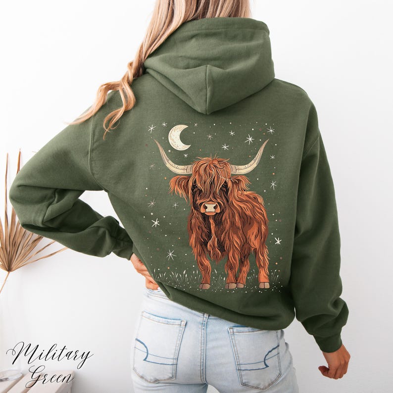 Op de afbeelding: Olijfgroene hoodie met een Highland-koeienafbeelding op de achterkant. Het ontwerp toont een bruine koe, een maan en sterren. De tekst "Military Green" staat linksonder.