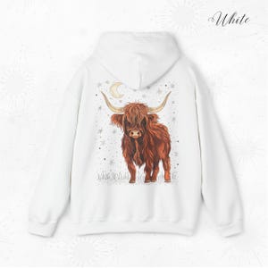Op de afbeelding: Witte hoodie met een gedetailleerde illustratie van een Schotse Hooglandkoe, met lange hoorns, tegen een achtergrond van sterren en een halve maan. Het woord "White" staat in cursief rechtsboven.