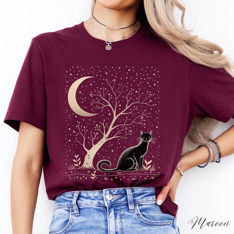 May include: Maroon t-shirt featuring a gold illustration of a crescent moon, tree, and stars, with a black cat sitting below. The design is set against a starry background. The word "Maroon" is written in script at the bottom right.
