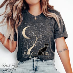 May include: Dark grey heather t-shirt featuring a gold crescent moon, tree, and stars design. A black cat sits beneath the tree. The text "Dark Grey Heather" is at the bottom of the shirt.