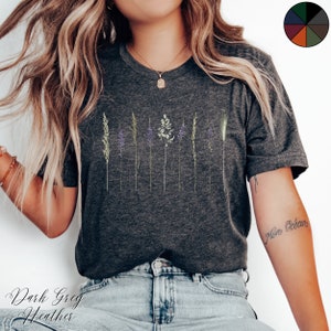 May include: A dark grey heather t-shirt featuring a graphic of a row of stylised grass and flowers. The shirt is being worn by a person with long blonde hair and blue jeans.