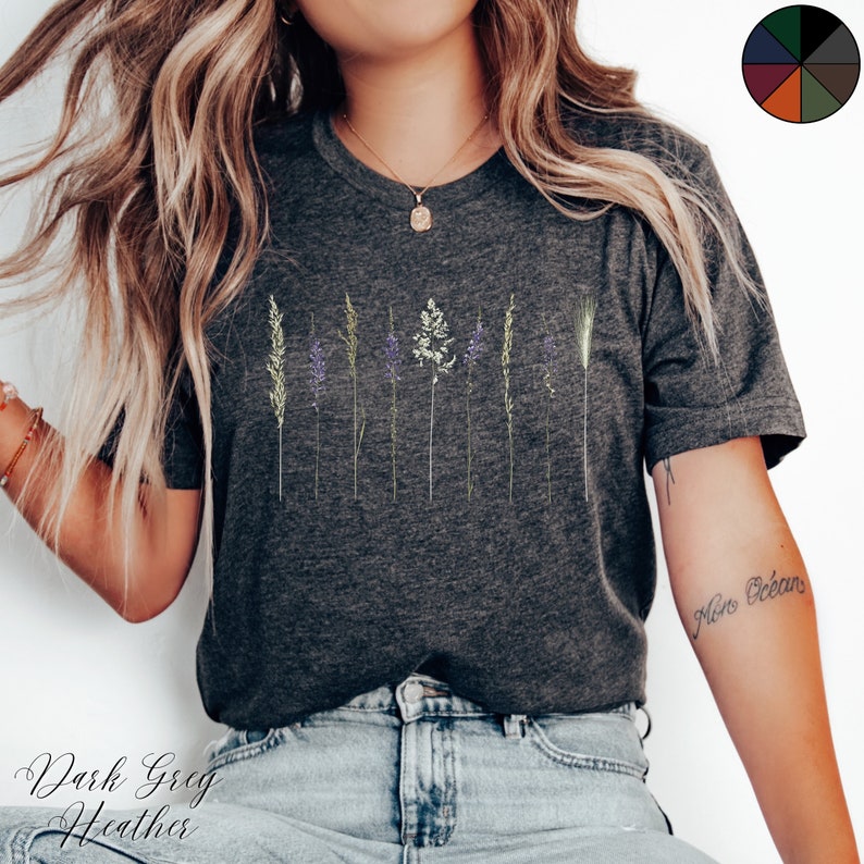 May include: A dark grey heather t-shirt featuring a graphic of a row of stylised grass and flowers. The shirt is being worn by a person with long blonde hair and blue jeans.