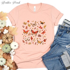 May include: A heather peach t-shirt with a graphic print of butterflies and fall leaves in shades of orange, red, and yellow.