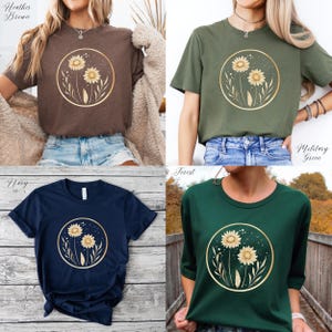 May include: Four t-shirts in different colors: heather brown, military green, navy, and forest green. Each shirt features a gold-colored circular design with two sunflowers and foliage. The shirts are short-sleeved and made of a soft material.