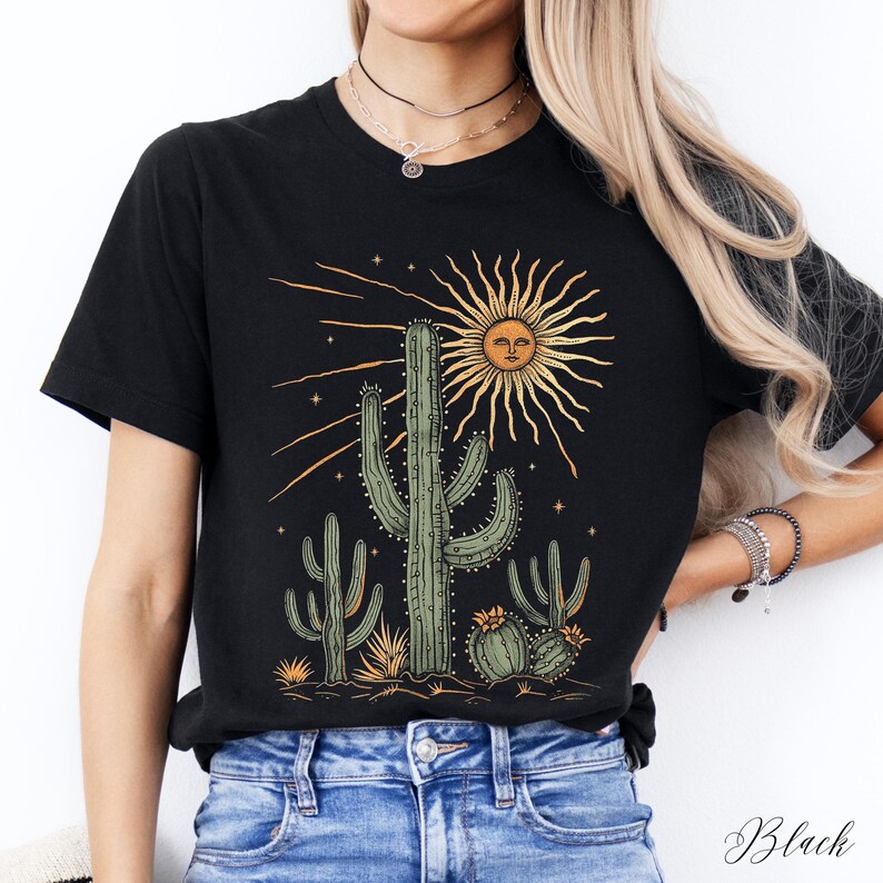 May include: Black t-shirt featuring a desert scene with cacti and a sun with a face. The graphic is in shades of green, yellow, and orange. The word "Black" is printed in cursive at the bottom right.