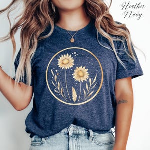 May include: Heather Navy t-shirt featuring a gold circular design with two sunflowers and leaves. The sunflowers have yellow petals and are surrounded by a gold outline. The text "Heather Navy" is written in cursive at the top right.