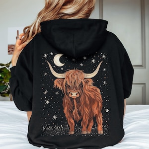 Op de afbeelding: Zwarte hoodie met een bruin Highland-koeienontwerp, een maan en sterren. De koe heeft lange hoorns en een ruige vacht. Het ontwerp staat tegen een donkere achtergrond, wat een hemels thema cre&euml;ert.