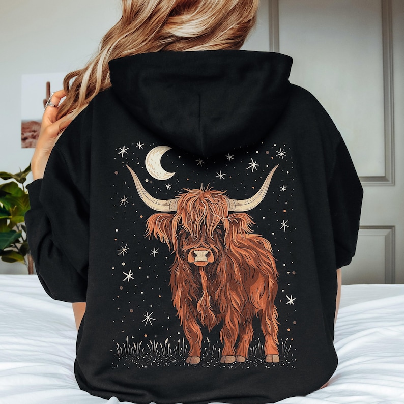 Op de afbeelding: Zwarte hoodie met een bruin Highland-koeienontwerp, een maan en sterren. De koe heeft lange hoorns en een ruige vacht. Het ontwerp staat tegen een donkere achtergrond, wat een hemels thema cre&euml;ert.