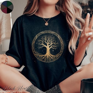 Può includere: Una maglietta nera con un design dorato ad albero della vita. L'albero è circondato da un cerchio con stelle e altri dettagli. Il design è intricato e dettagliato.