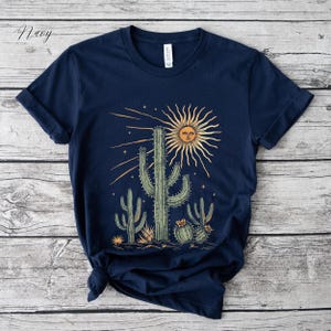 May include: Navy blue t-shirt featuring a desert scene with cacti and a smiling sun graphic. The design includes various cacti in shades of green and yellow, with a radiant sun and stars. The shirt is made of a soft material.