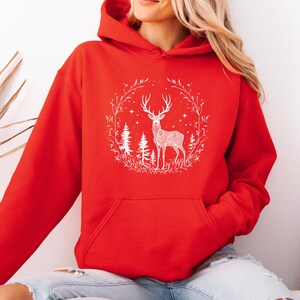 May include: A red hooded sweatshirt featuring a white graphic of a deer inside a circular wreath with trees and stars. The sweatshirt has a kangaroo pocket and a drawstring hood. The design is centered on the front of the garment.