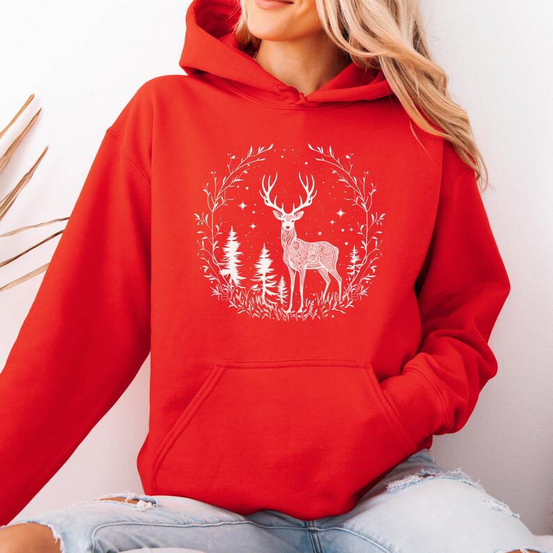 May include: A red hooded sweatshirt featuring a white graphic of a deer inside a circular wreath with trees and stars. The sweatshirt has a kangaroo pocket and a drawstring hood. The design is centered on the front of the garment.