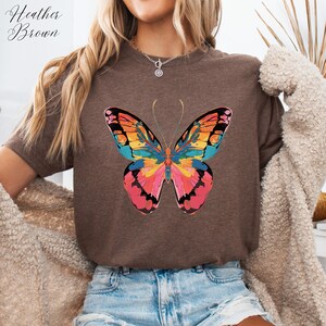 Pu&ograve; includere: T-shirt marrone 'Heather Brown' con un design a farfalla colorato. La farfalla ha ali rosa, arancioni, gialle e blu. La maglietta &egrave; di colore marrone neutro.