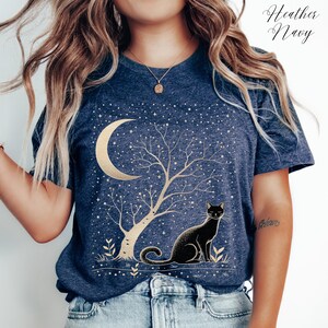May include: Heather Navy t-shirt featuring a gold crescent moon, tree, and stars design with a black cat. The shirt has a crew neck and short sleeves. The text "Heather Navy" is in the upper right corner.