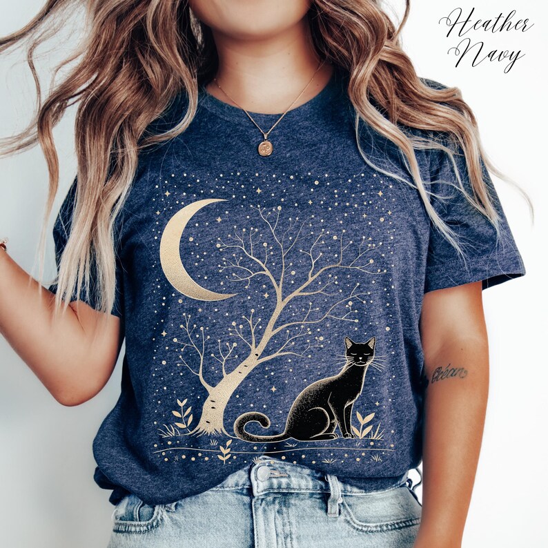 May include: Heather Navy t-shirt featuring a gold crescent moon, tree, and stars design with a black cat. The shirt has a crew neck and short sleeves. The text "Heather Navy" is in the upper right corner.