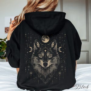 Peut inclure: Sweat-shirt à capuche noir avec un imprimé graphique d'une tête de loup avec un troisième œil et une lune croissante de chaque côté. Le loup est entouré d'étoiles et d'autres symboles célestes.