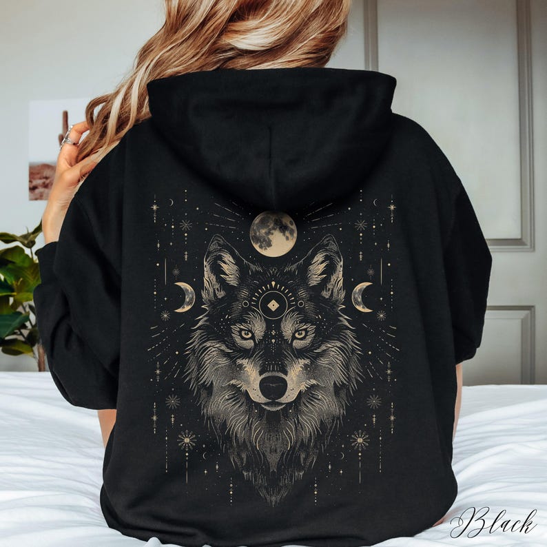 K&ouml;nnte beinhalten: Schwarzer Kapuzenpullover mit einem grafischen Aufdruck eines Wolfskopfes mit einem dritten Auge und einer Mondsichel auf jeder Seite. Der Wolf ist von Sternen und anderen himmlischen Symbolen umgeben.