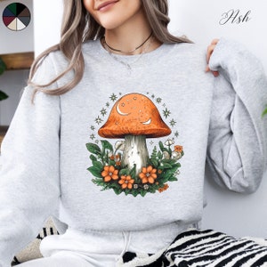 Puede incluir: Sudadera gris claro con un diseño de hongo. El hongo tiene un sombrero naranja con detalles de luna y estrellas, rodeado de hojas verdes y flores naranjas. La palabra "Ash" está impresa en la parte superior derecha.